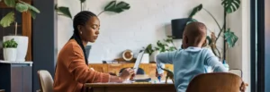 A mother and her child sitting at a table in a home setting while the mother is working, representing an empathetic approach to wellbeing at work and hybrid working.