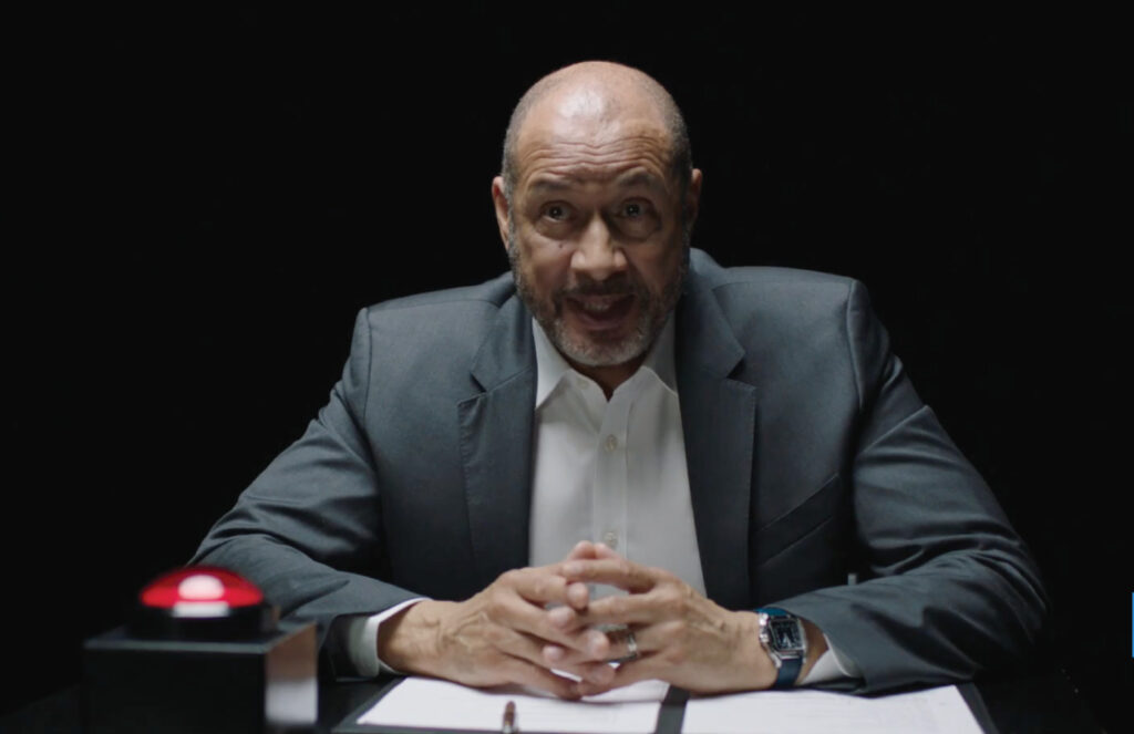 A man in a suit sitting at an interview table in the dark. On the table in front of him are pieces of paper sprawled across the table and a big red button.